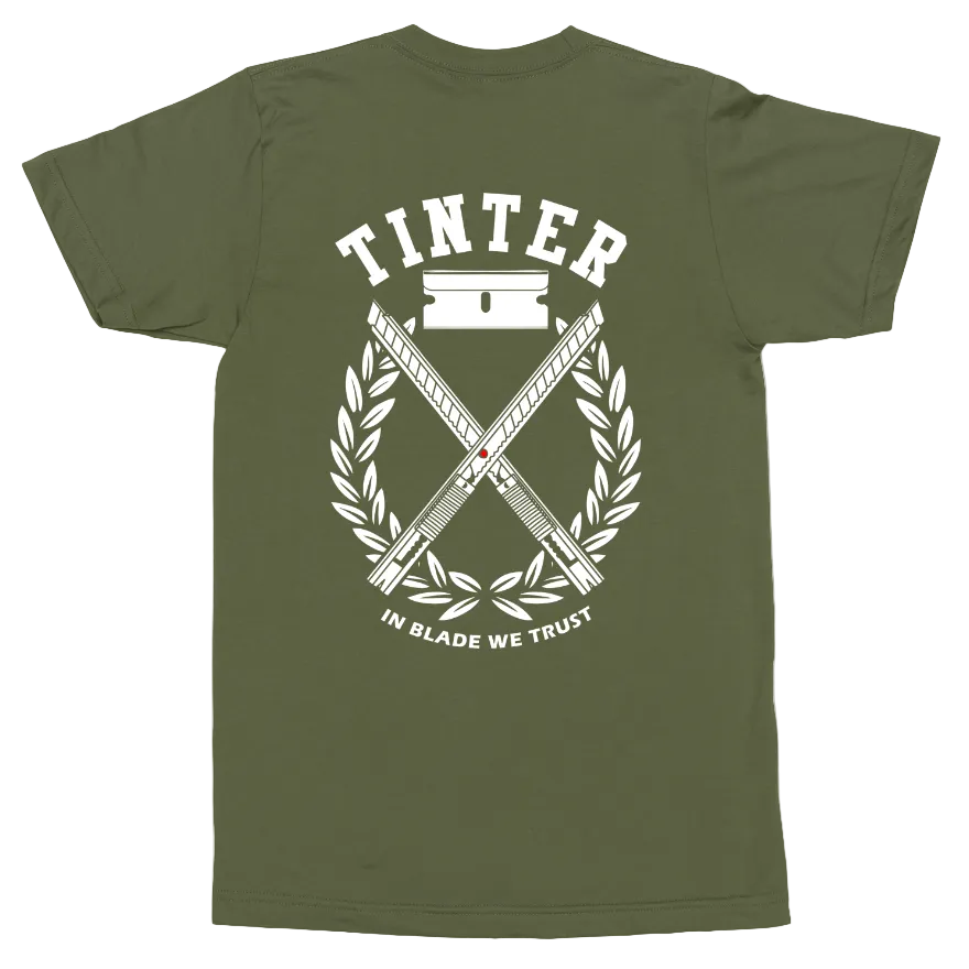 An olive green short-sleeve t-shirt with a white graphic print on the back. The graphic features the brand name 'TINTER' above crossed swords, a laurel wreath, and the motto 'IN BLADE WE TRUST'.