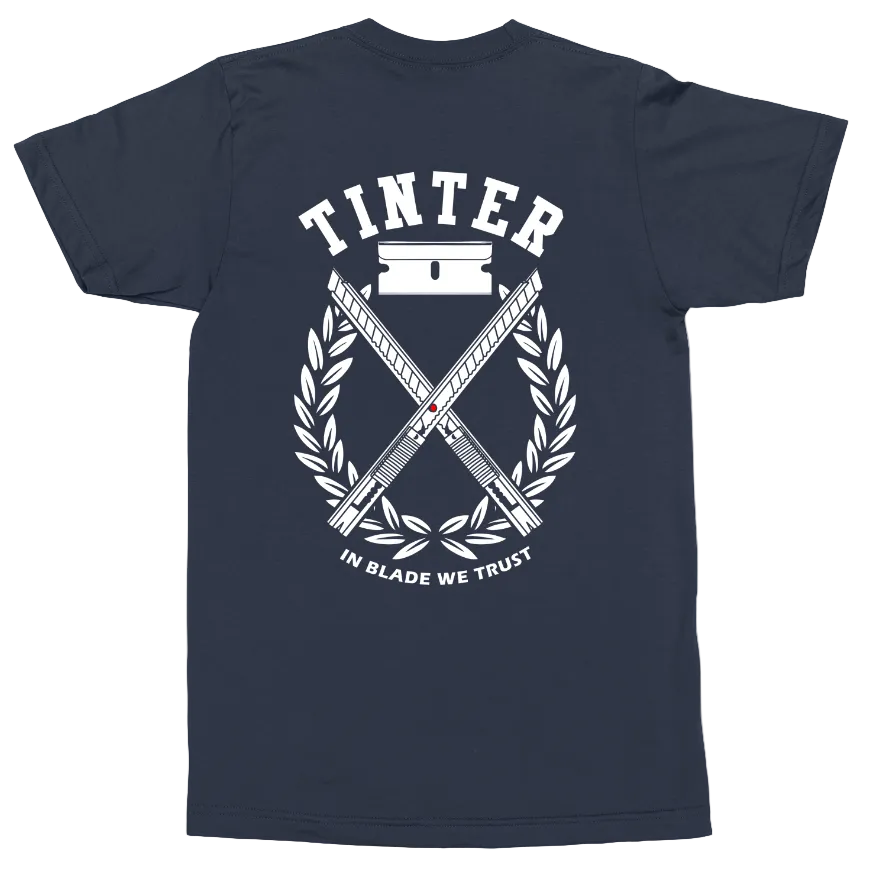 A navy blue short-sleeve t-shirt shown from the back, featuring a large white graphic print with the word 'TINTER' above crossed blades, a laurel wreath, and the phrase 'IN BLADE WE TRUST'.