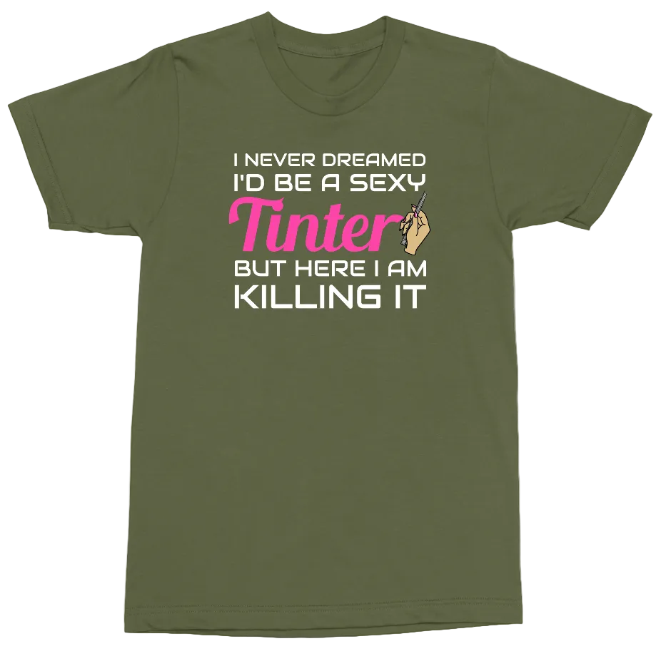 An olive green short-sleeve t-shirt with a graphic text print on the front reading 'I NEVER DREAMED I'D BE A SEXY Tinter BUT HERE I AM KILLING IT' in white and pink, accompanied by a small hand graphic.