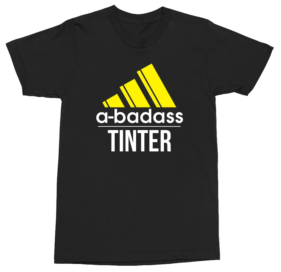 A black short-sleeve t-shirt with a graphic print featuring a yellow Adidas-style logo, the text 'a-badass' in white, and 'TINTER' in white below it, shown on a plain white background.