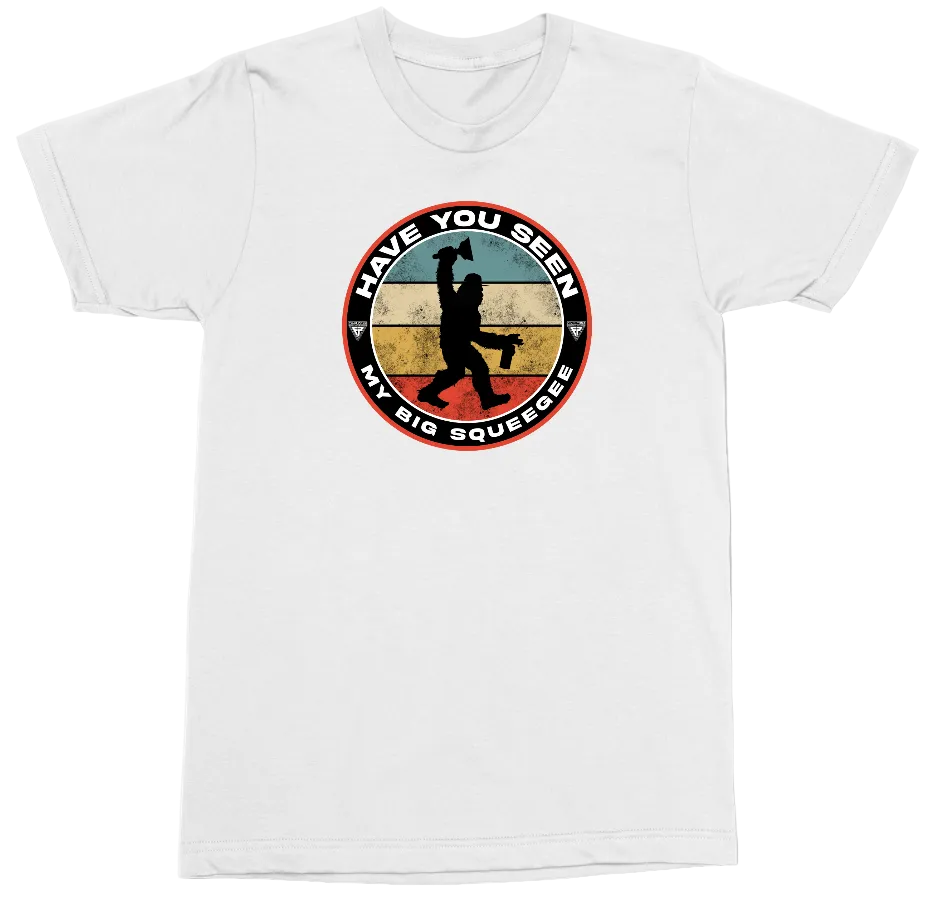 A white short-sleeve crewneck t-shirt featuring a circular graphic on the chest. The graphic shows a black silhouette of a person doing a squintz (holding a penis in the vagina) against a distressed background of blue, yellow, and red stripes, with the text 'HAVE YOU SEEN MY BIG SQUEEEZER' in white lettering.
